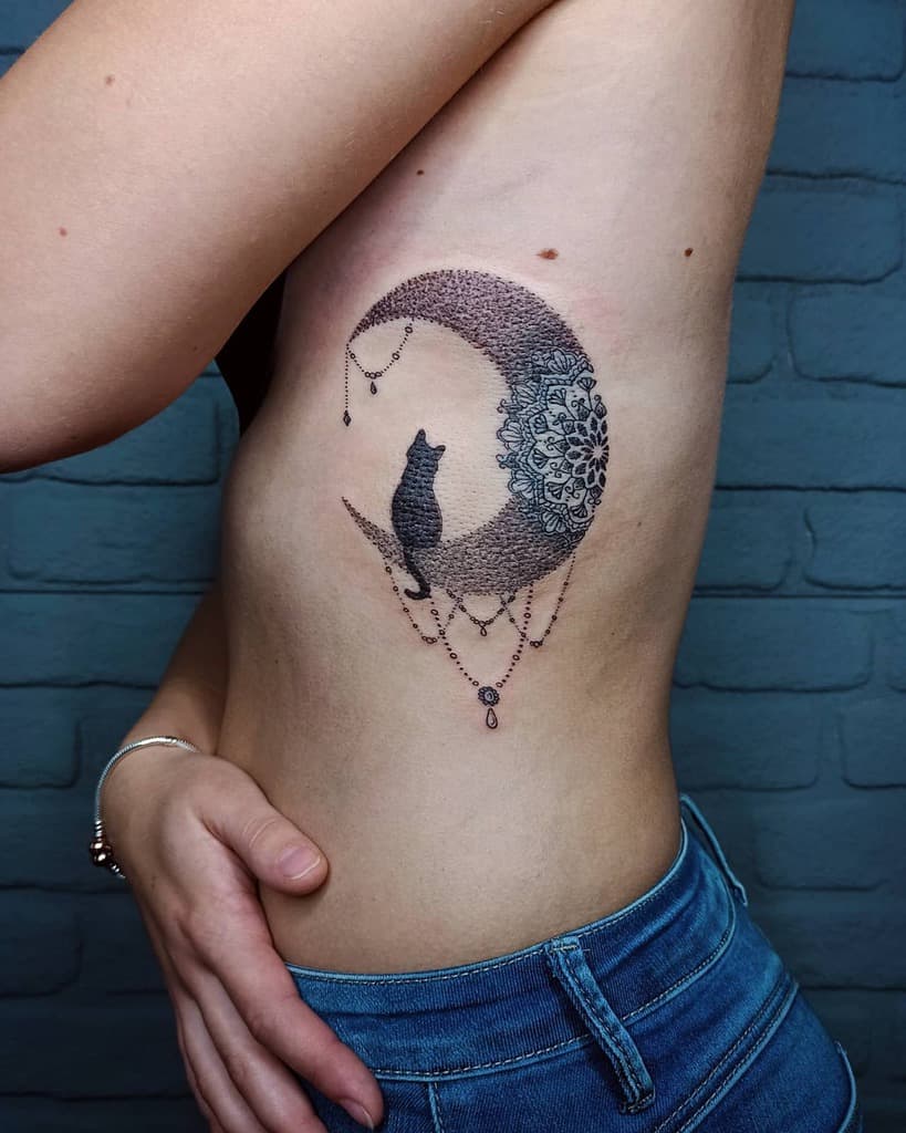 A side tattoo of a cat perched on a crescent moon, adorned with intricate designs and dangling chains, elegantly graces the person's rib