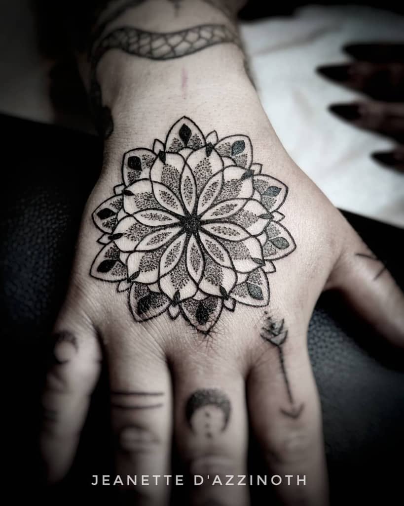 A detailed geometric mandala flower tattoo, surrounded by other small tattoos such as arrow, dots, crescent moon and lines