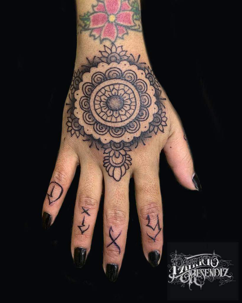 Ornate mandala tattoo on a hand, with symbols on fingers and a pink flower tattoo above the wrist