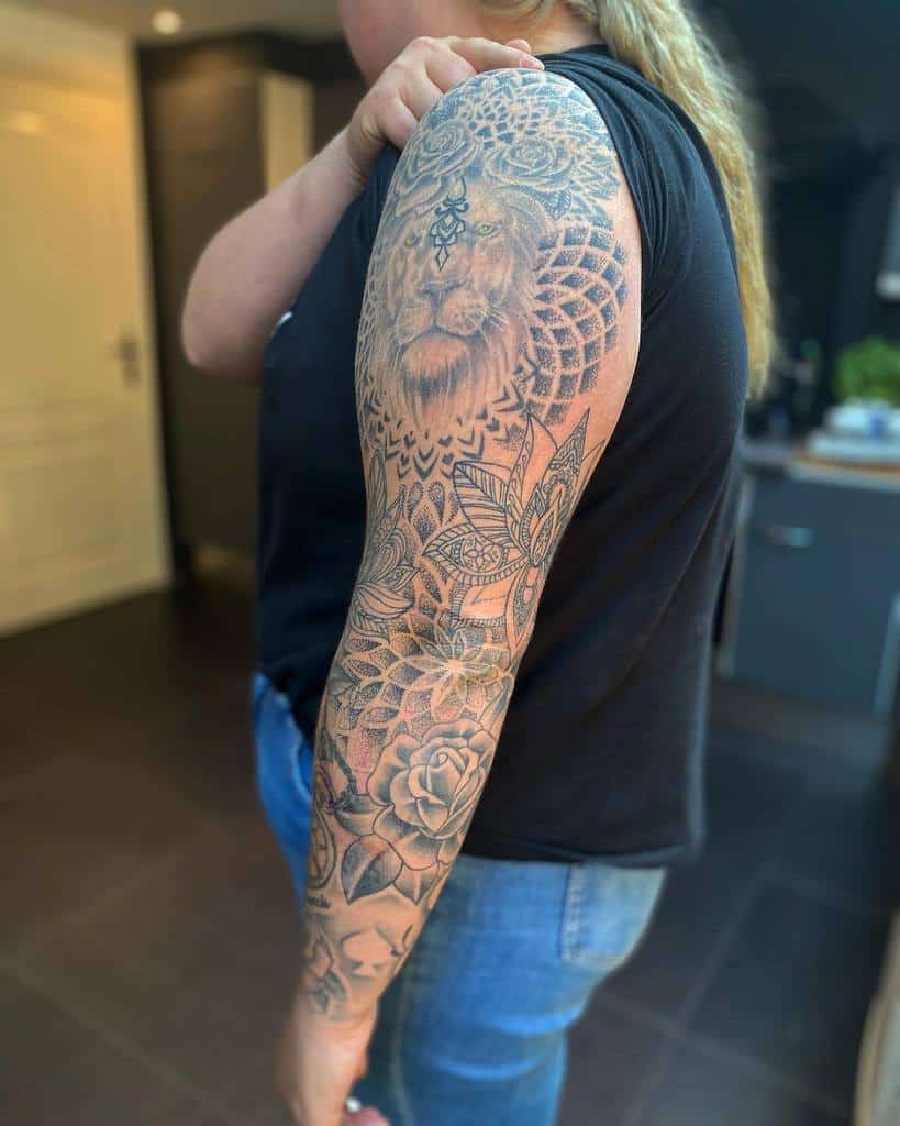 A black and grey full sleeve tattoo featuring a detailed lion's face on the upper arm, surrounded by mandala patterns, flowers, and intricate geometric designs extending down to the wrist