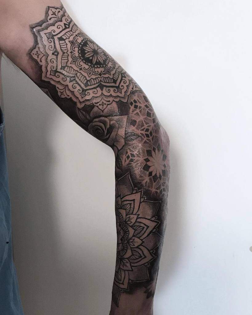 A black and grey sleeve tattoo featuring intricate mandala patterns, geometric shapes, and shading, extending from the shoulder down the arm with a rose incorporated into the design