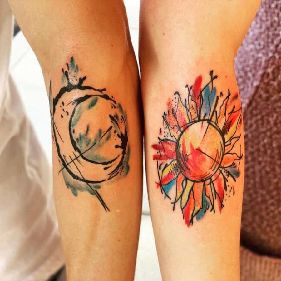 Abstract sun and moon tattoos on forearms, featuring bold black ink strokes with watercolor-style blue, red, and yellow accents.