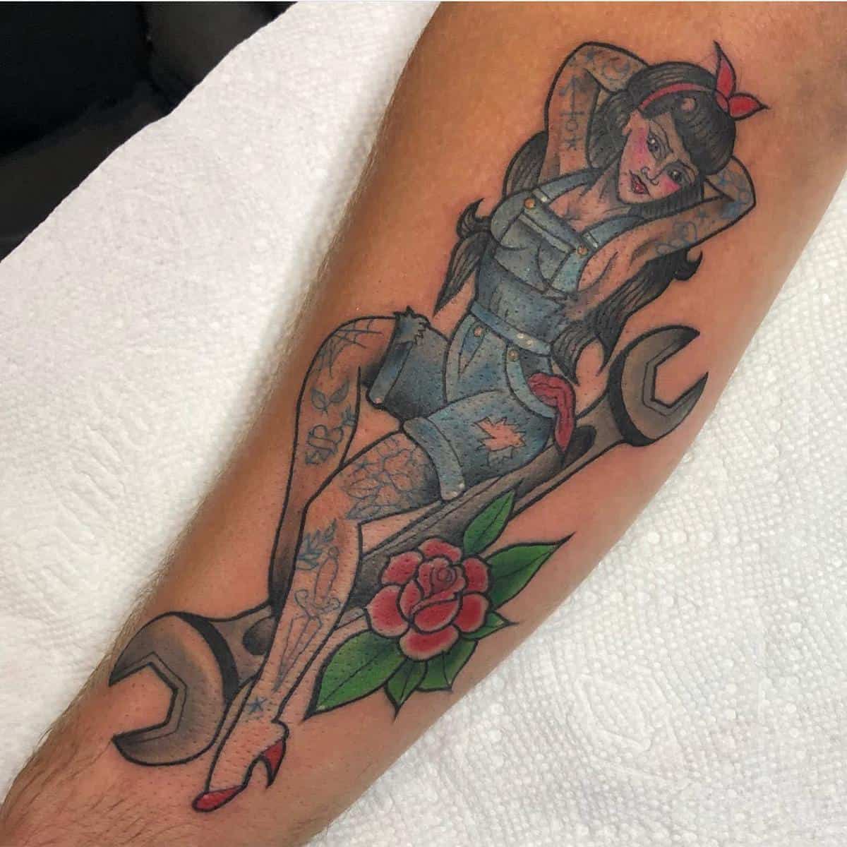 Tattoo on an arm featuring a pin-up girl in denim sitting on a wrench with a red rose and green leaves below her