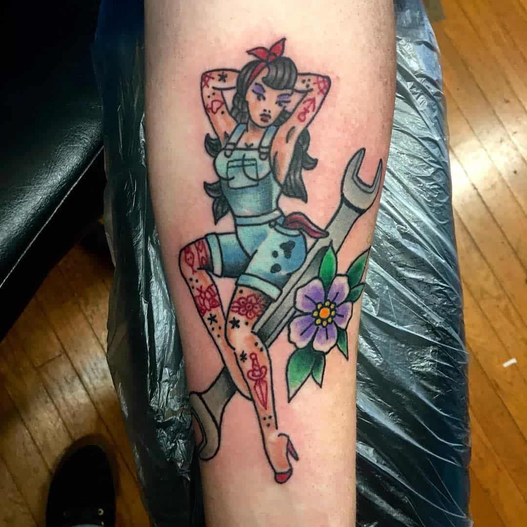 Tattoo of a pin-up girl with tattoos sitting on a wrench, accompanied by a flower and leaves on a forearm