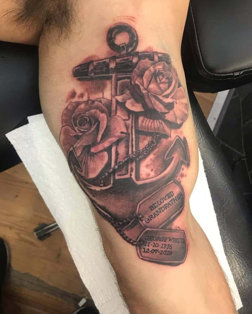 Tattoo of a navy anchor adorned with roses and a ribbon reading "Beloved Grandfather" along with dates