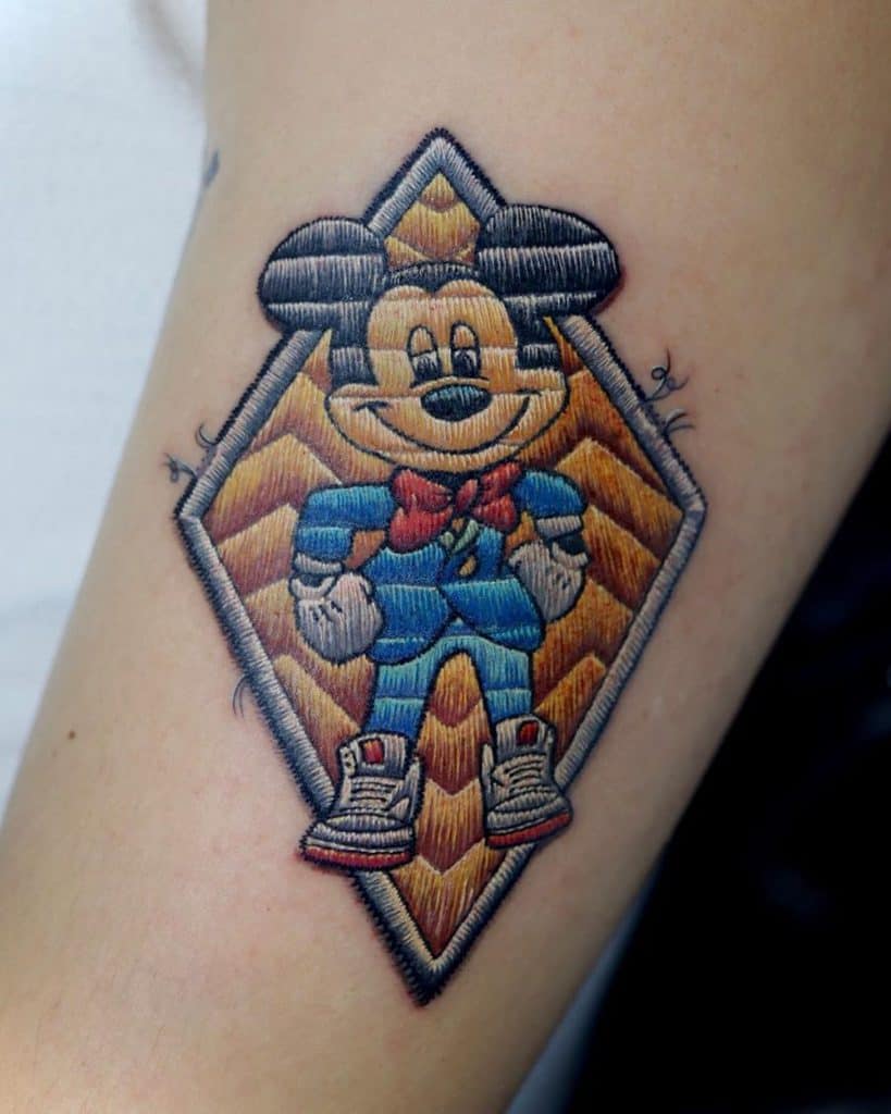 Textile-effect tattoo of Mickey Mouse in modern attire, with detailed stitching and quilted shading for a fabric-inspired design.