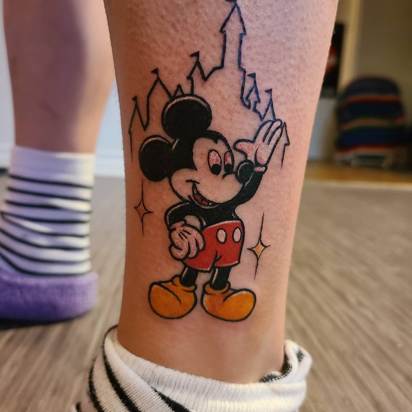 Colorful tattoo of Mickey Mouse waving, with a castle outline in the background on an ankle