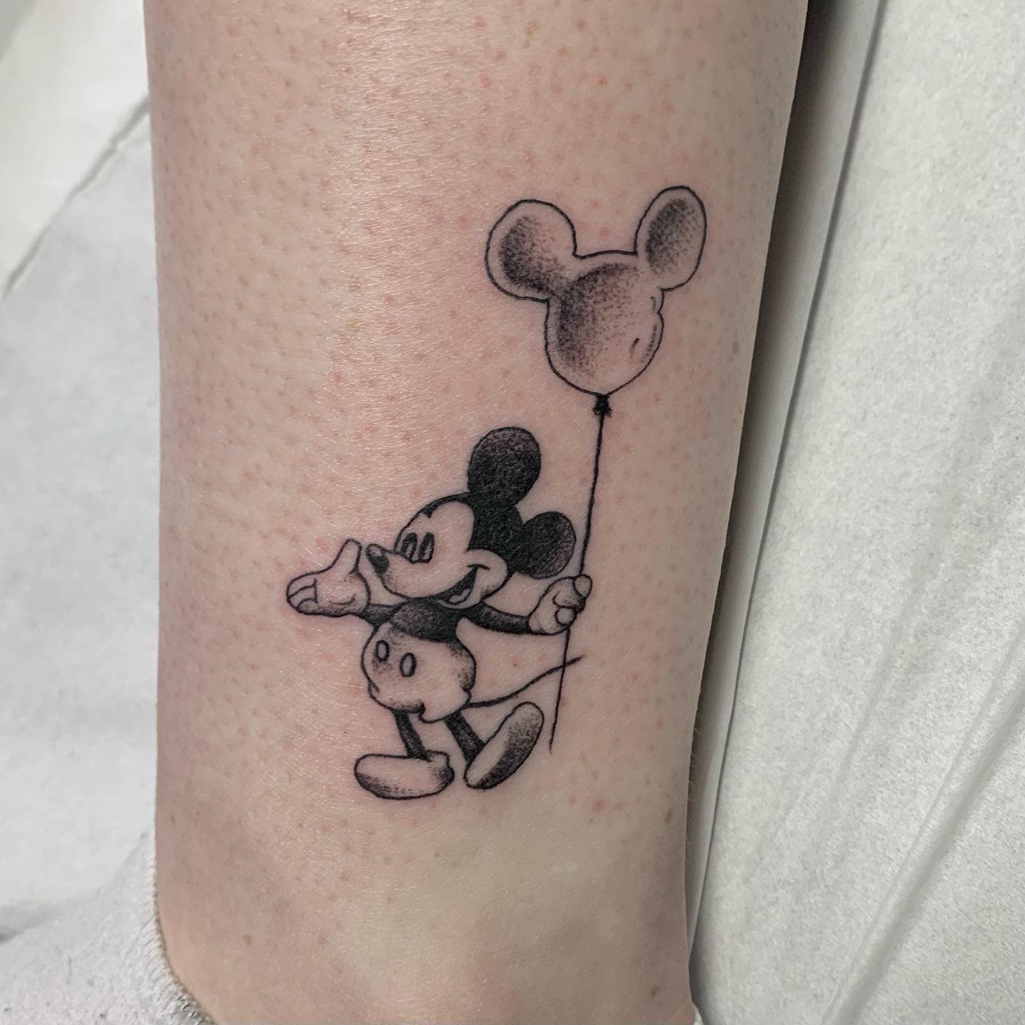 Black-and-gray tattoo of Mickey Mouse holding a Mickey-shaped balloon on an ankle