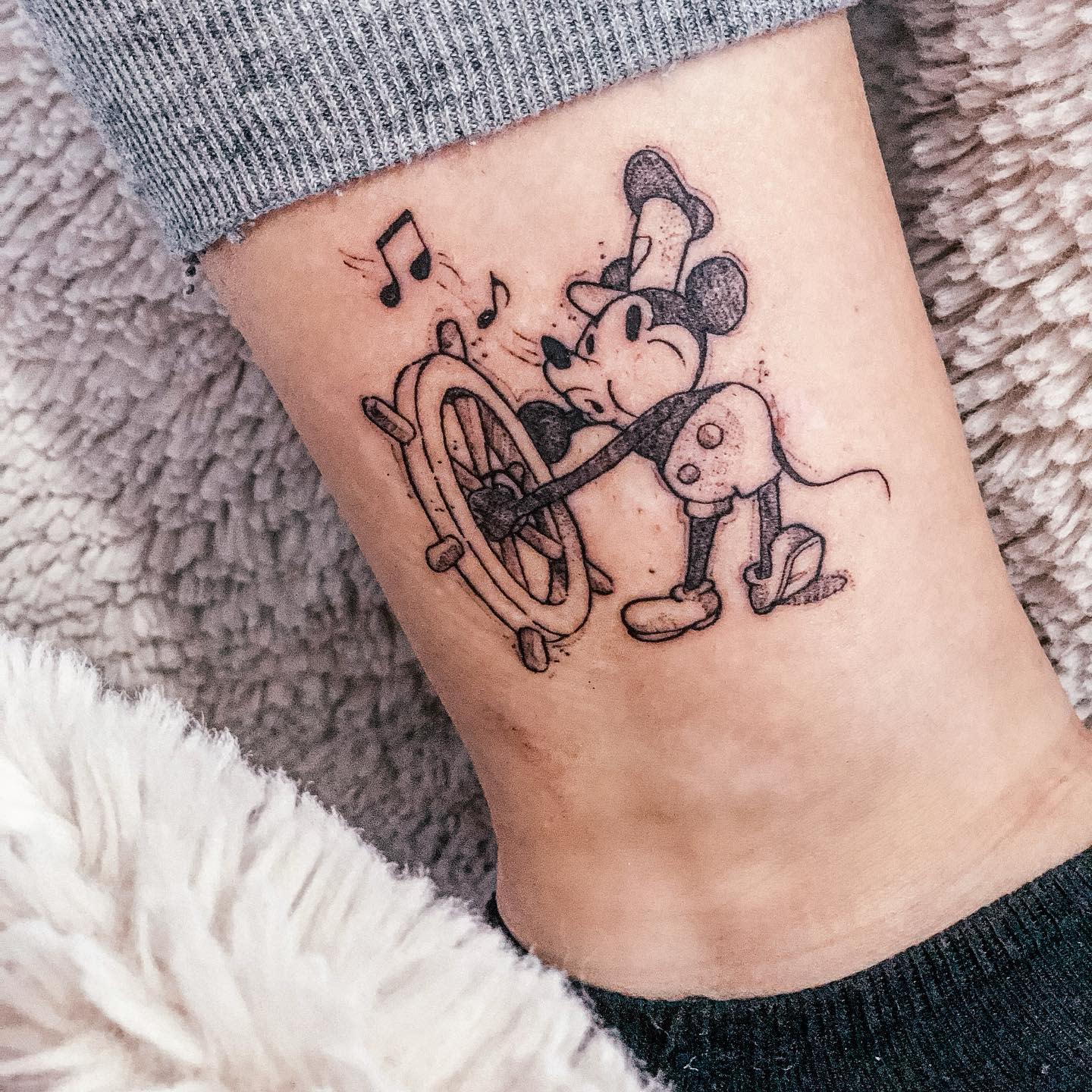 Black-and-gray tattoo of Steamboat Willie Mickey Mouse steering a ship's wheel on an ankle