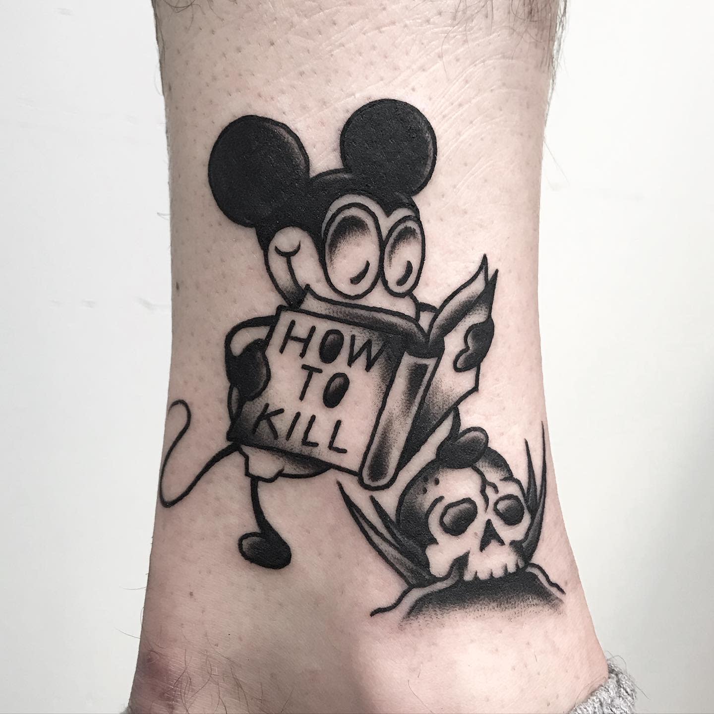Black-and-gray tattoo of a Mickey Mouse figure reading a book titled 'How to Kill' next to a skull
