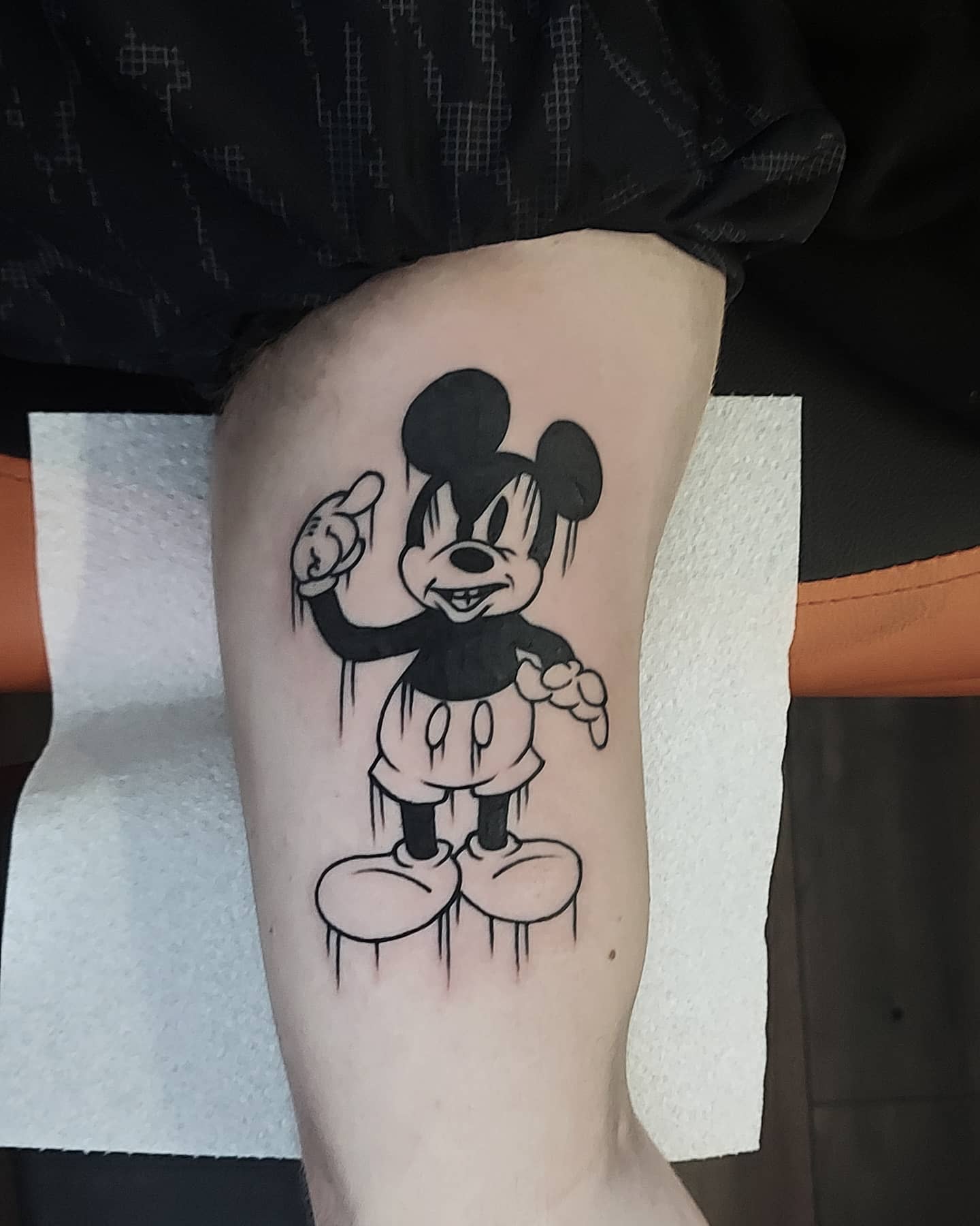 Black-and-gray tattoo of a creepy Mickey Mouse with exaggerated eyes and sharp teeth, pointing upwards