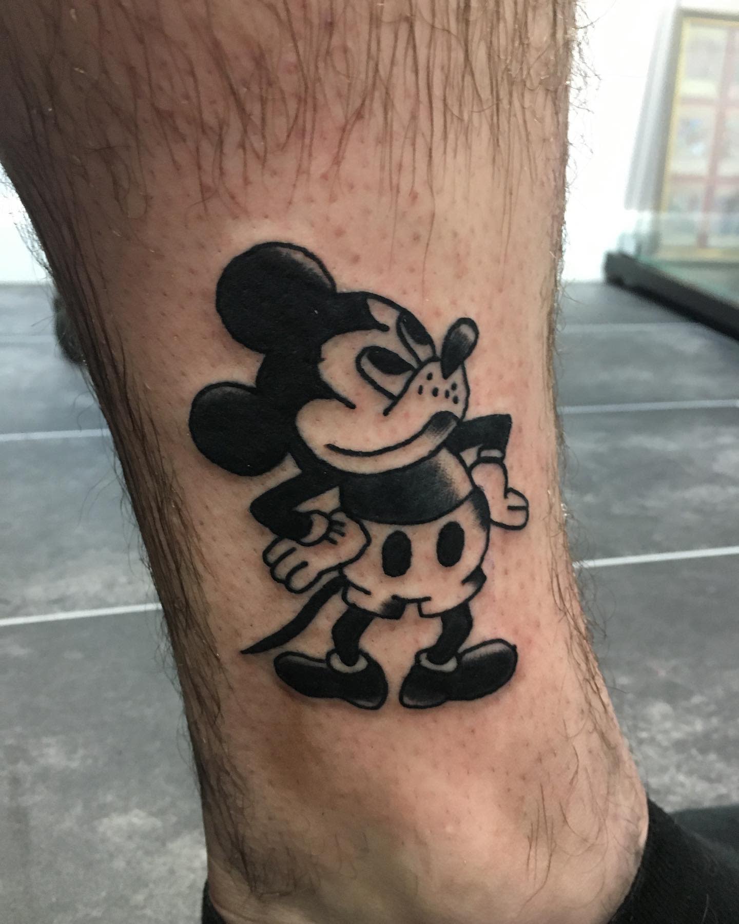 Bold black-and-gray tattoo of an angry-looking Mickey Mouse with arms on hips, placed on the ankle