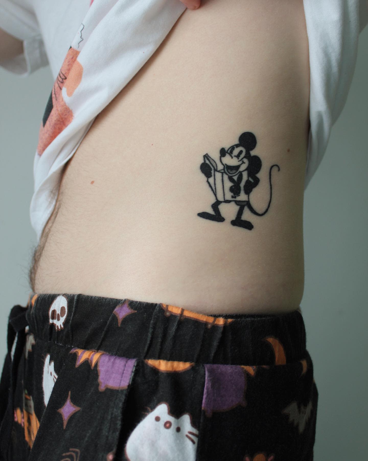 Small black-and-gray tattoo of a Mickey Mouse figure reading a book on the side of the torso