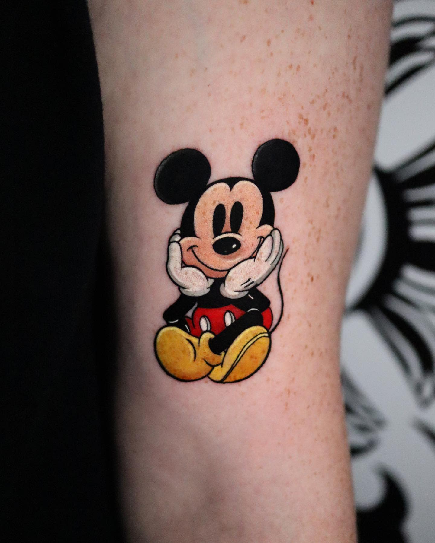 Colorful tattoo of a cute Mickey Mouse sitting with his chin resting on his hands, smiling