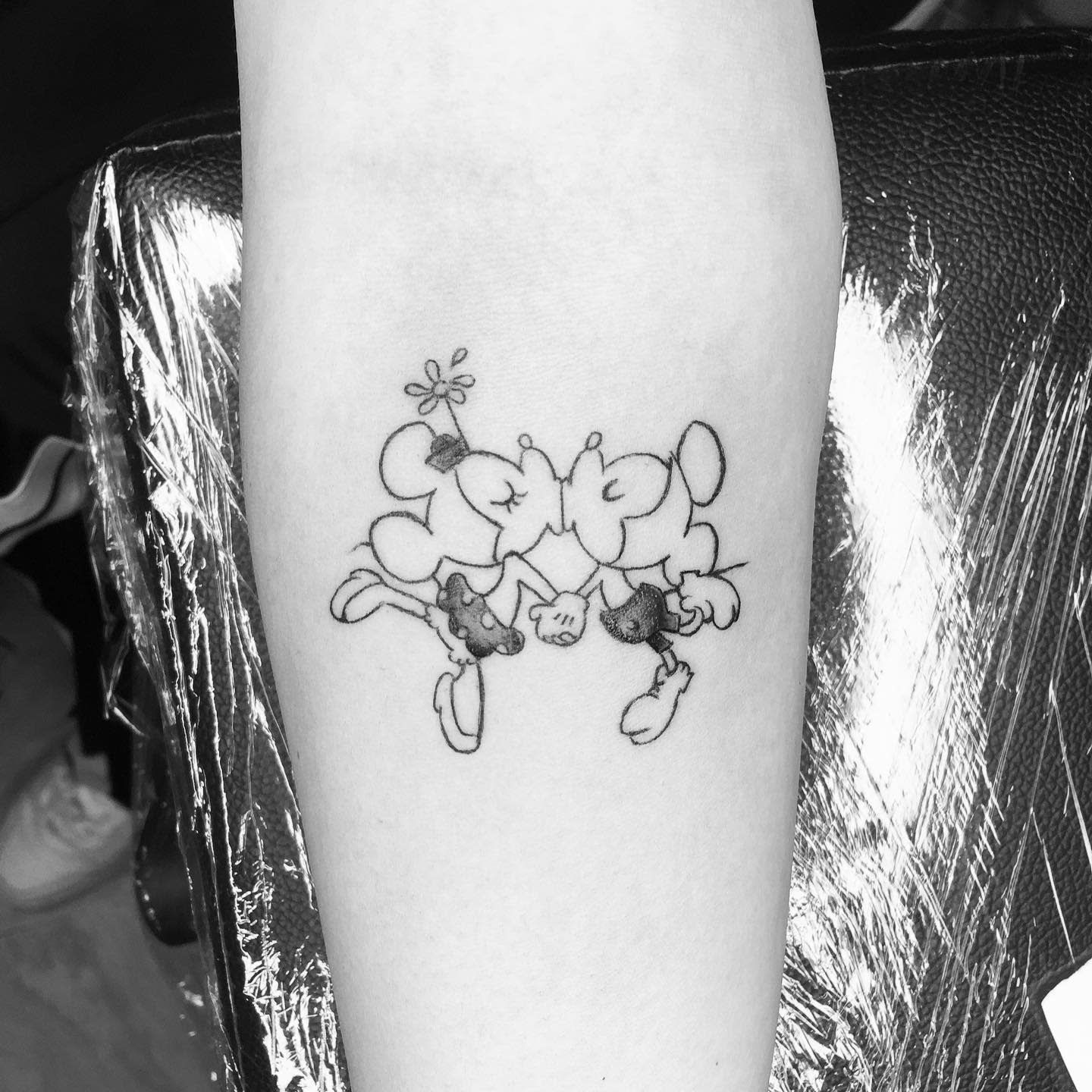 Simple black-and-gray tattoo of Mickey and Minnie Mouse kissing, with their noses forming a heart shape