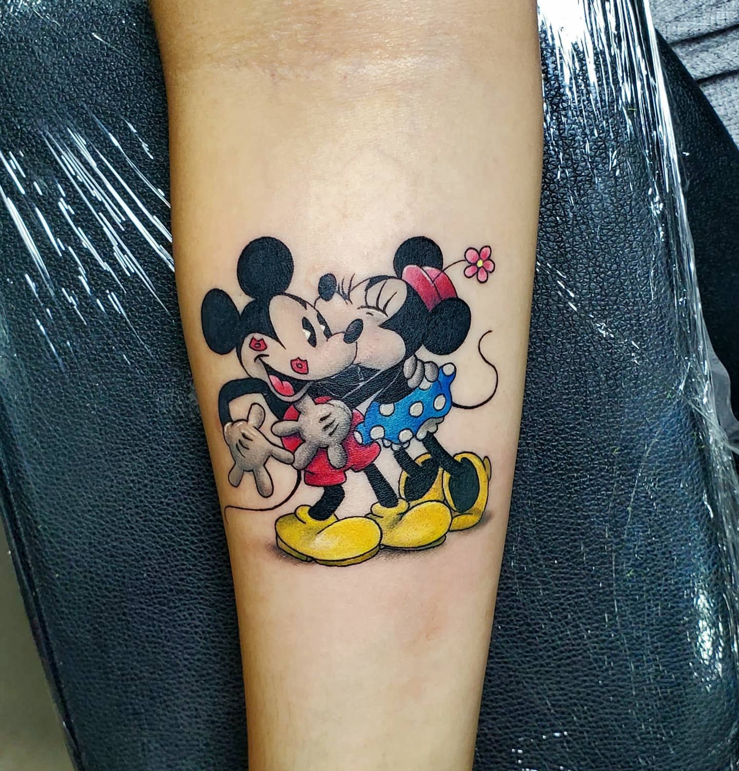 Colorful tattoo of Mickey and Minnie Mouse hugging and kissing, with Minnie wearing a blue polka dot skirt