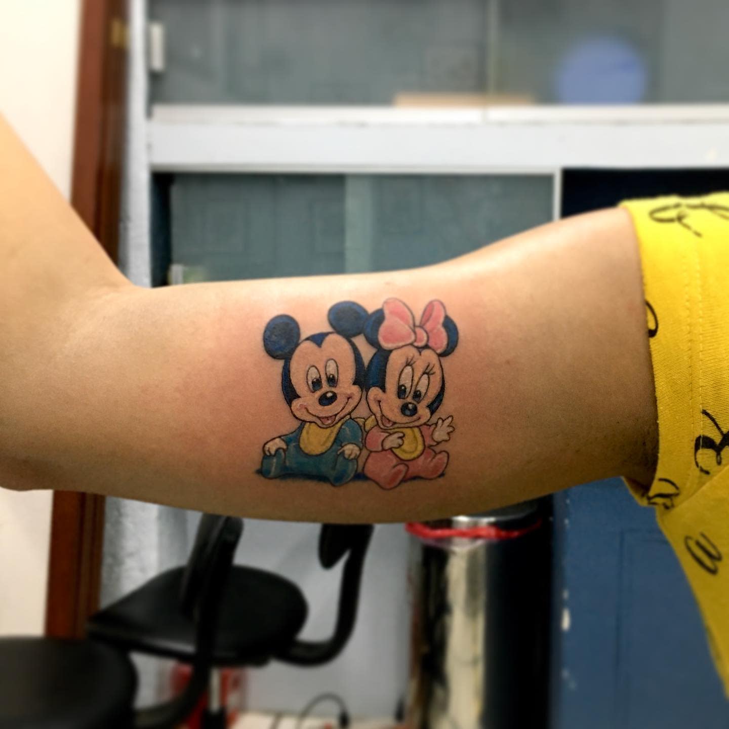 Colorful tattoo of baby Mickey and Minnie Mouse sitting side by side, with Minnie wearing a pink bow