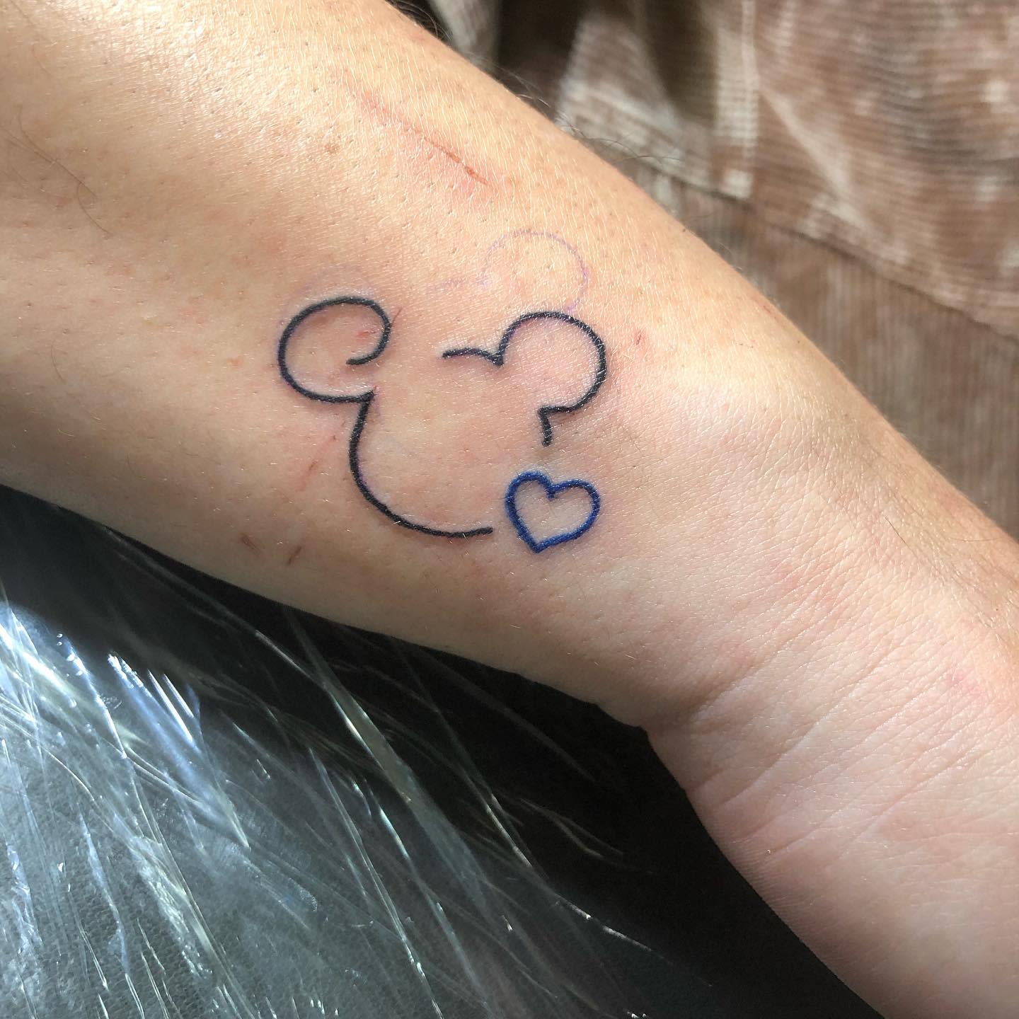 Simple tattoo of an outline of Mickey Mouse's head with a small heart shape below it