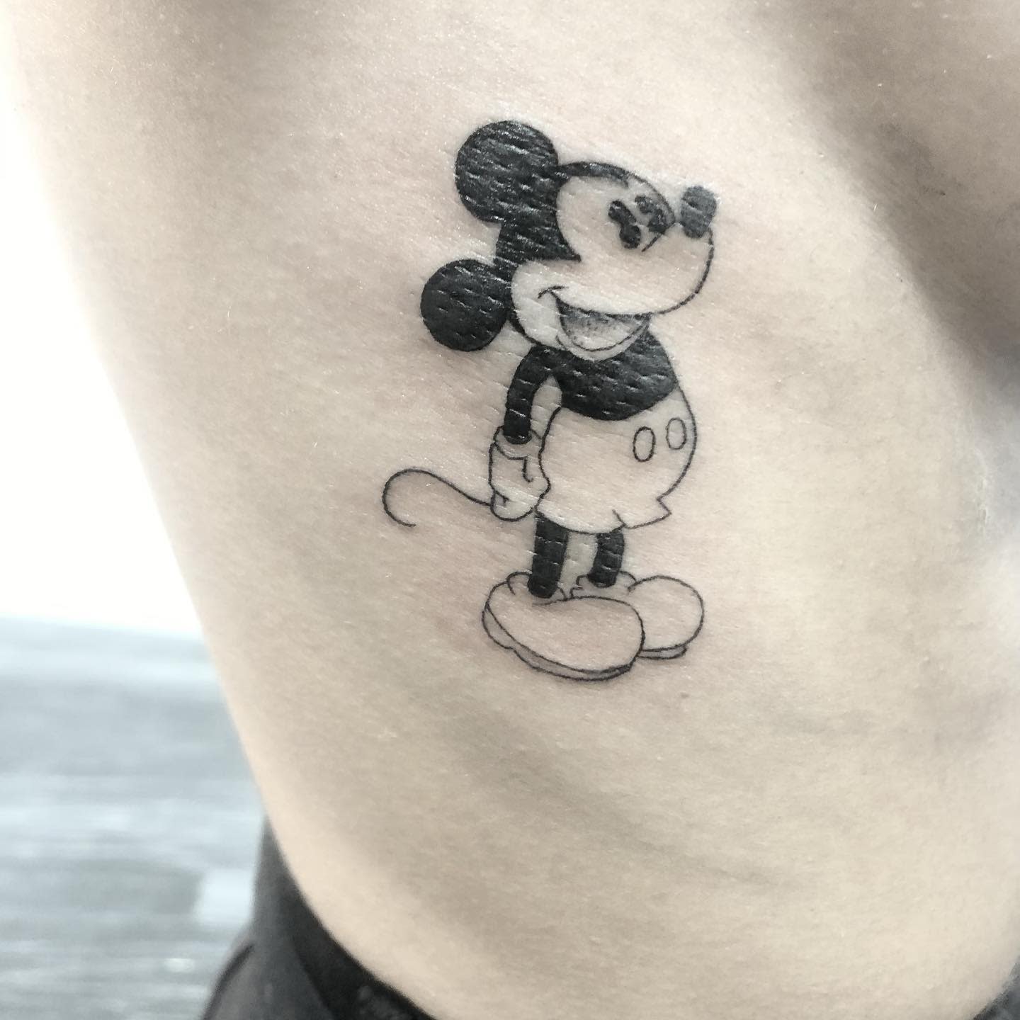 Small black-and-gray tattoo of classic Mickey Mouse standing with a smile, placed on the ribcage