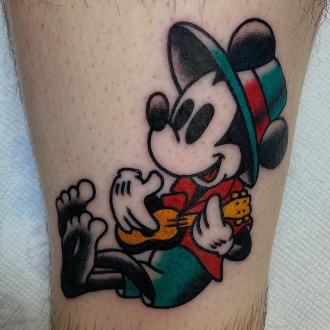 Colorful tattoo of Mickey Mouse dressed in a hat and jacket, playing a small guitar, with a traditional tattoo style