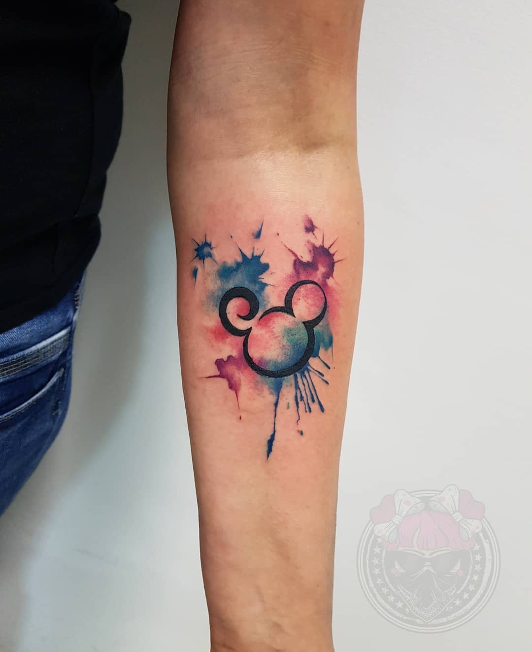 Watercolor tattoo of Mickey Mouse's head silhouette with splashes of blue and pink on the forearm