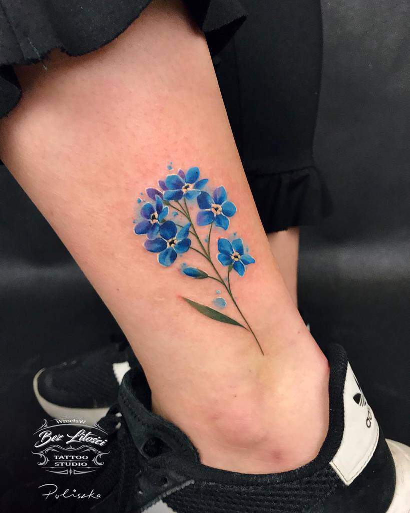 A tattoo of vibrant blue flowers with green stems on the side of an ankle