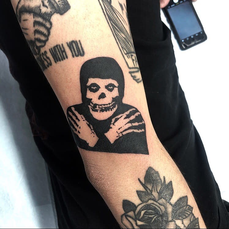 Black tattoo of a skeleton figure crossing its arms on an upper arm
