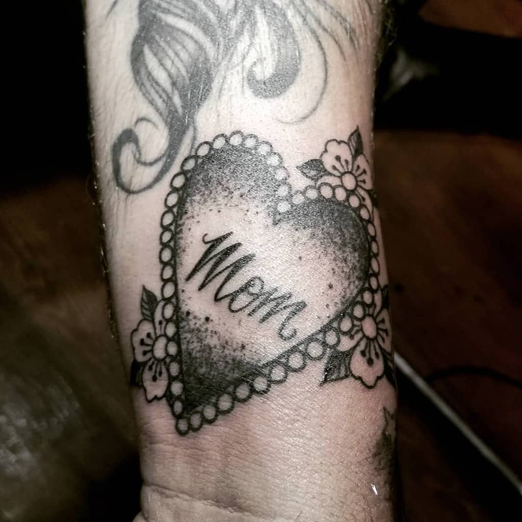 Heart-shaped tattoo with the word 'Mom' in elegant script, adorned with small flowers on an arm