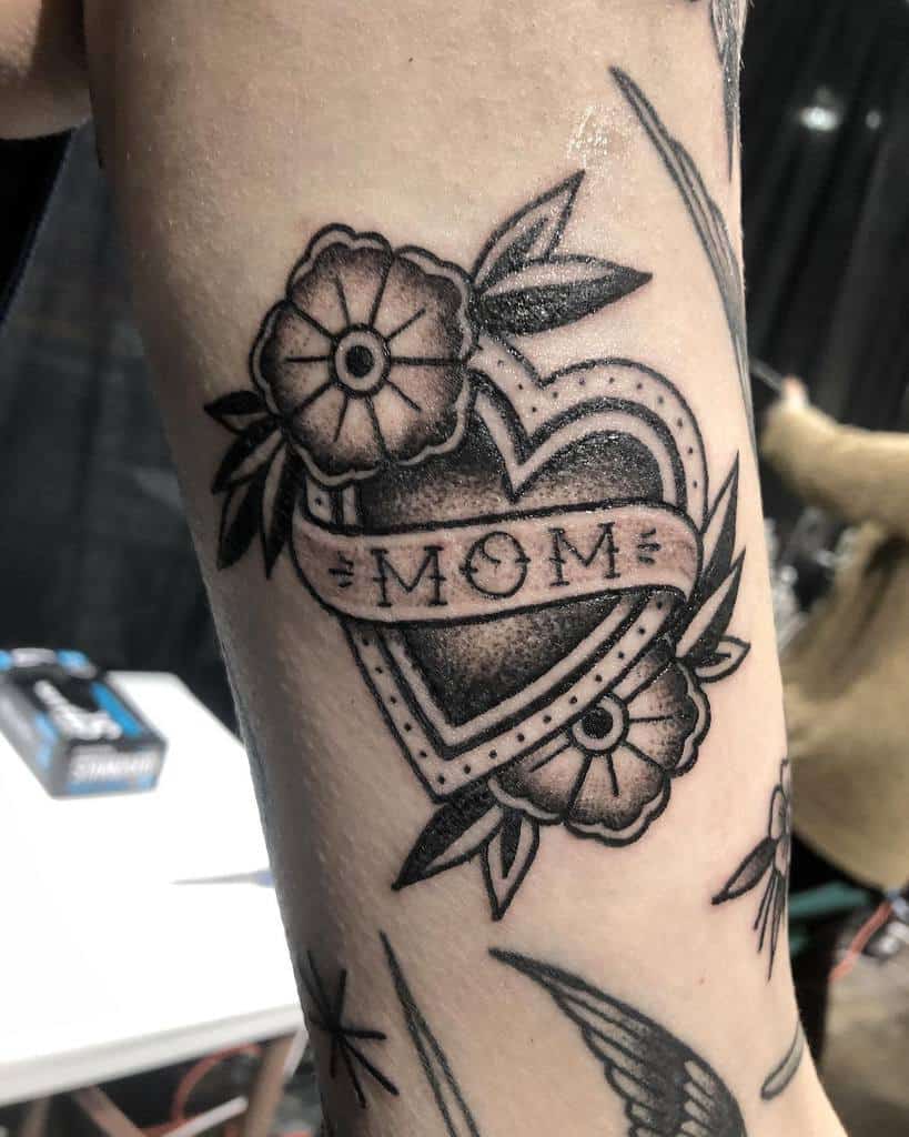 Tattoo of a heart with a "MOM" banner and flowers beautifully inked on an arm