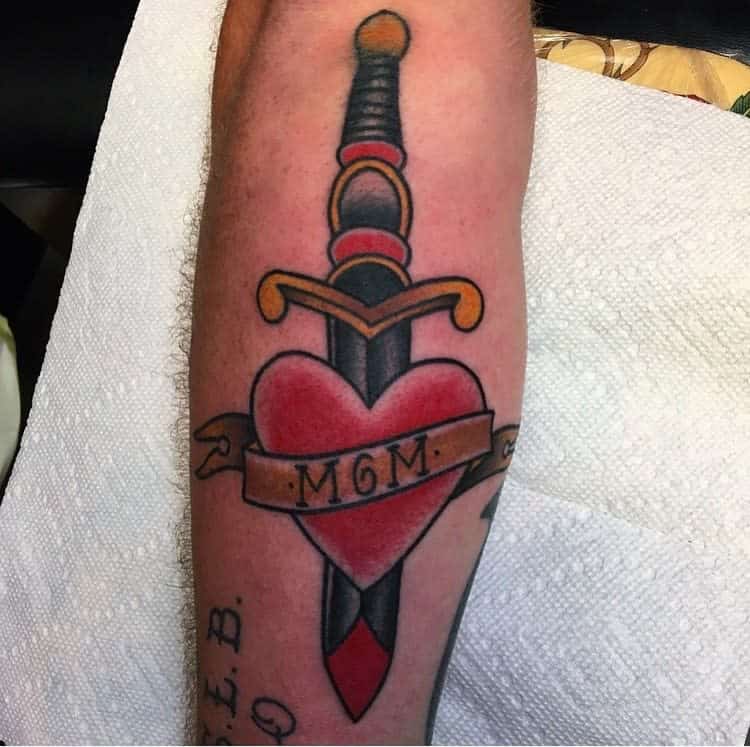 Tattoo on an arm of a dagger piercing a red heart with a "MOM" banner elegantly wrapped around it