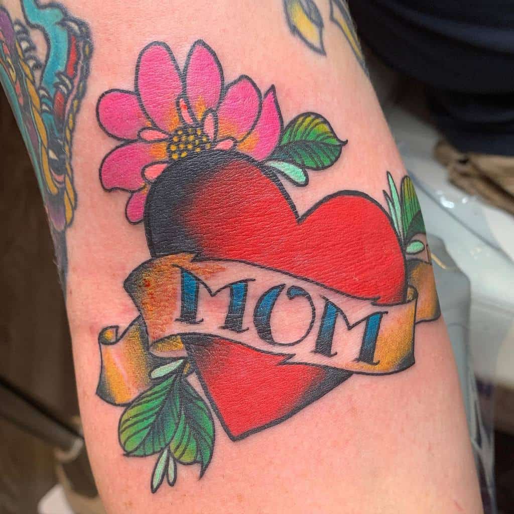 Tattoo of a heart with a "MOM" banner, adorned with a pink flower and lush green leaves