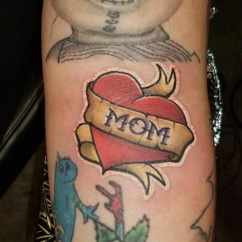 Tattoo of a red heart with a yellow banner reading "MOM," seamlessly nestled among other intricate ink designs on an arm