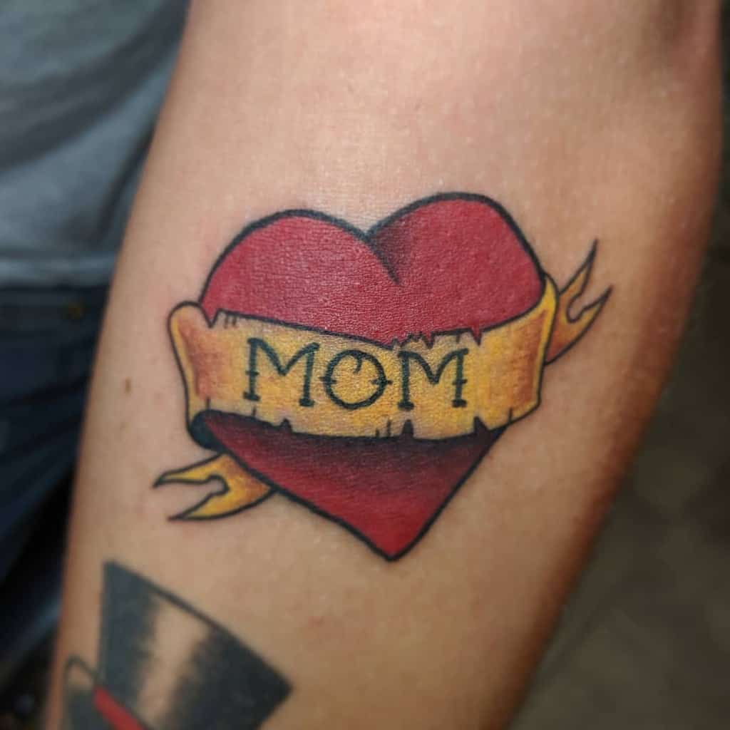 A vibrant tattoo of a red heart with a yellow banner reading "MOM" adorns the person's arm