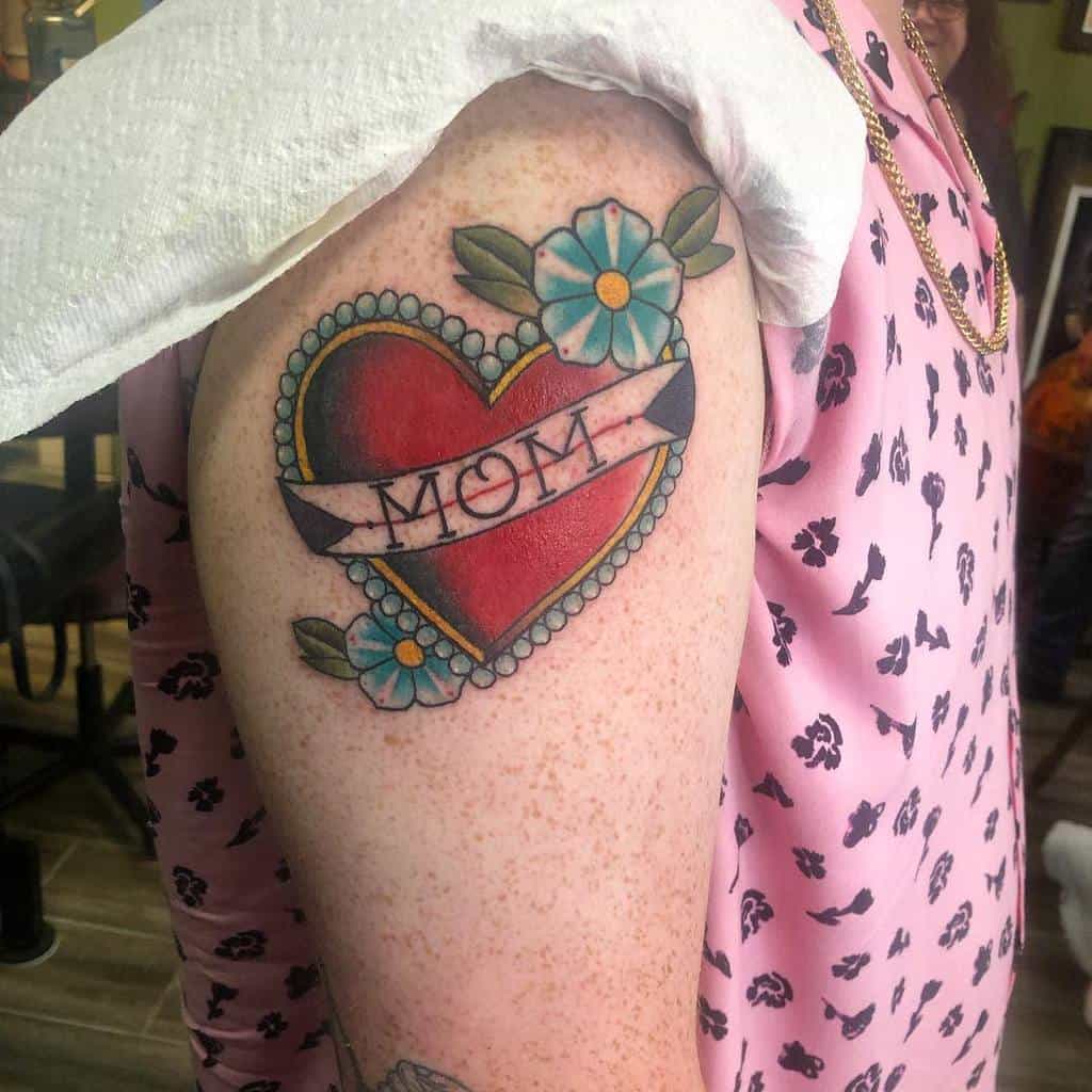Tattoo of a heart with a 'MOM' banner and blooming flowers elegantly inked on a person's upper arm