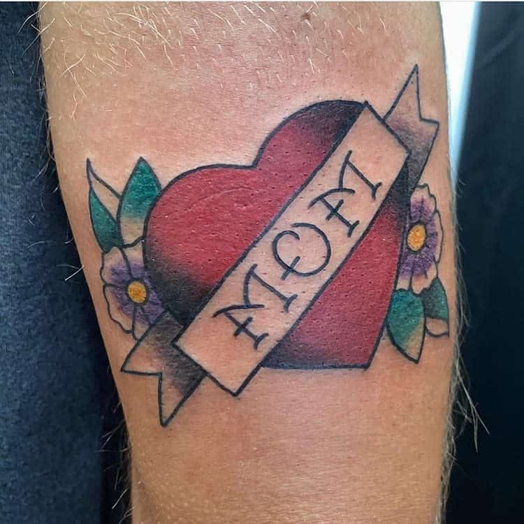 Tattoo of a heart with a banner reading "Mom," beautifully surrounded by flowers and vibrant green leaves