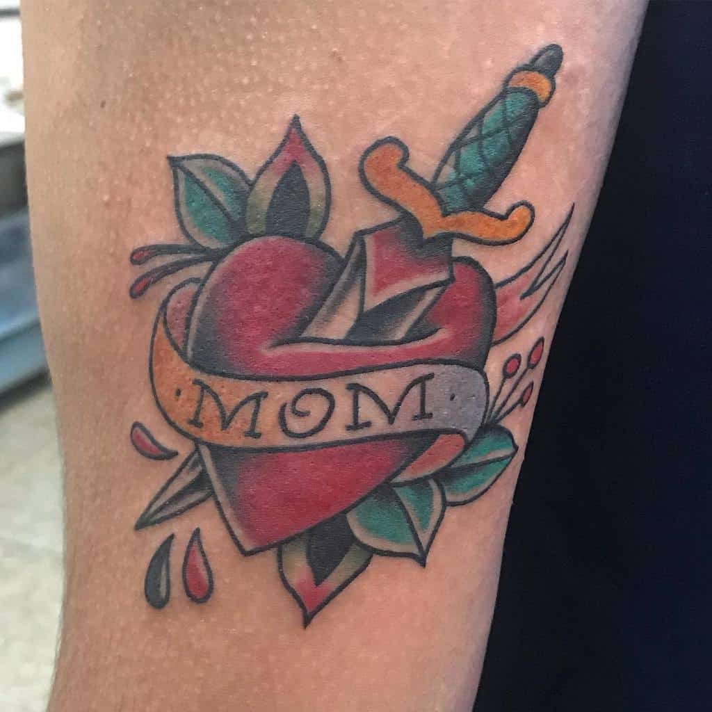 Tattoo of a heart with "MOM" written on a banner, elegantly pierced by a dagger with drops of blood