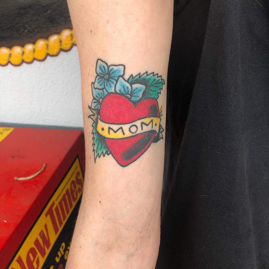 Tattoo of a heart with "MOM" in a banner, surrounded by blue flowers and green leaves