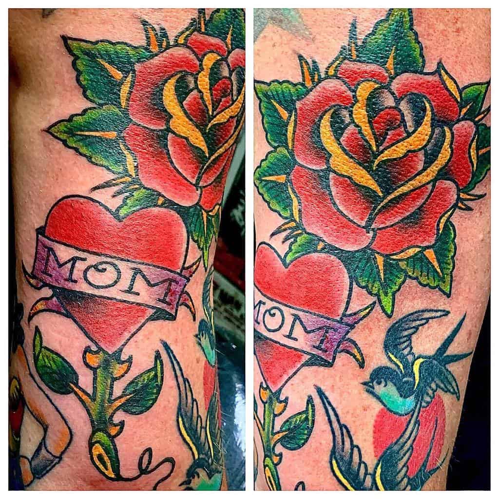 Tattoo of a red heart with a "MOM" banner, rose, and a green bird gracefully adorning a person's arm