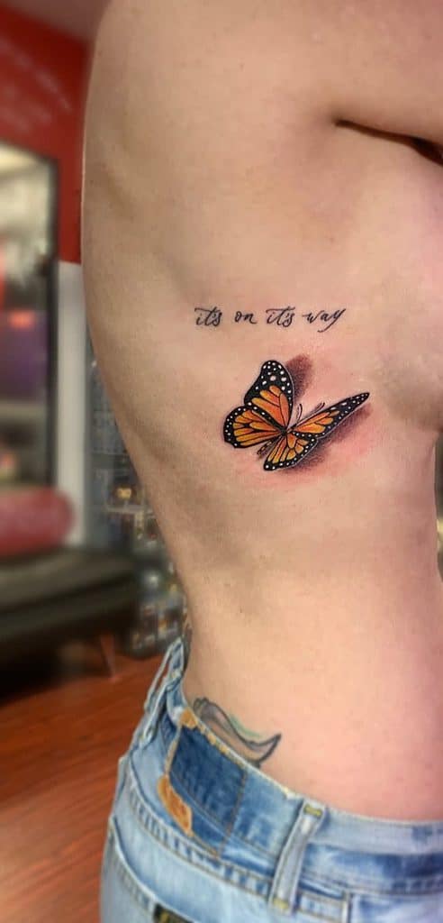 A monarch tattoo features a butterfly beside the cursive text "it's on its way" on a womans side