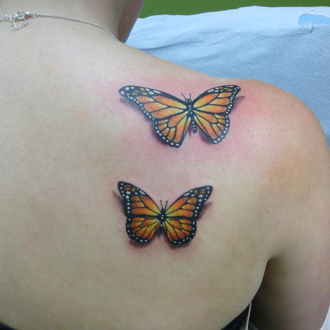 Two realistic monarch butterfly tattoos in vibrant orange and black adorn a person's upper back and shoulder