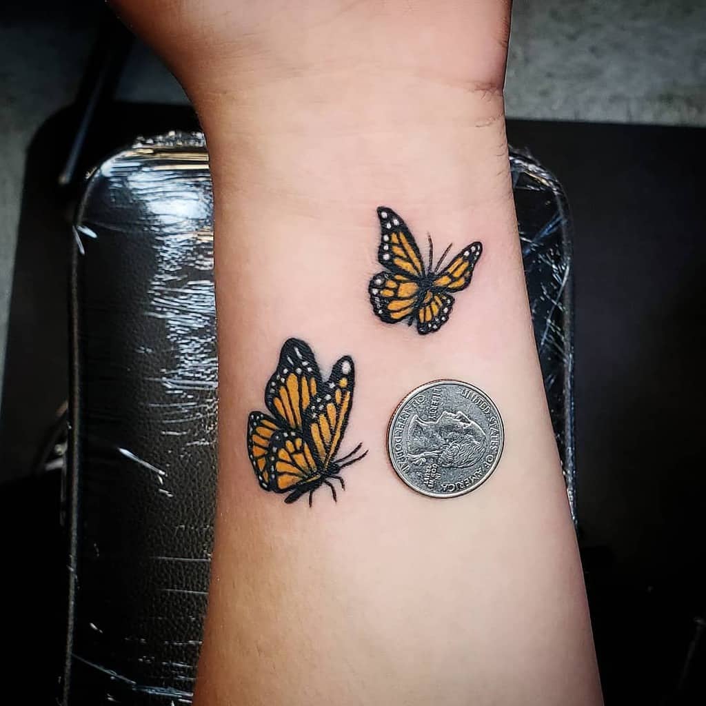 Two small monarch butterfly tattoos on the wrist, featuring vibrant orange and black details, placed beside a quarter for size comparison.