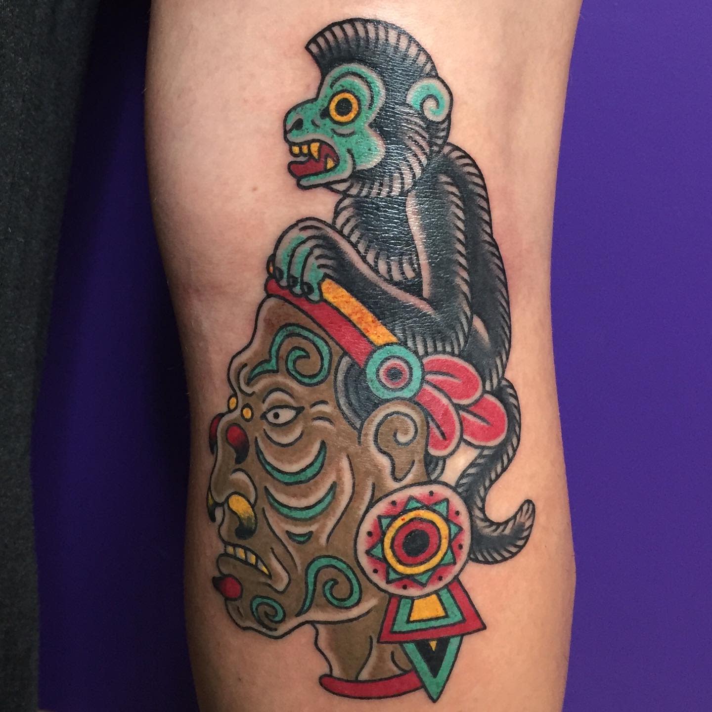 Tattoo of a colorful monkey sitting on a stylized, Aztec-inspired human head