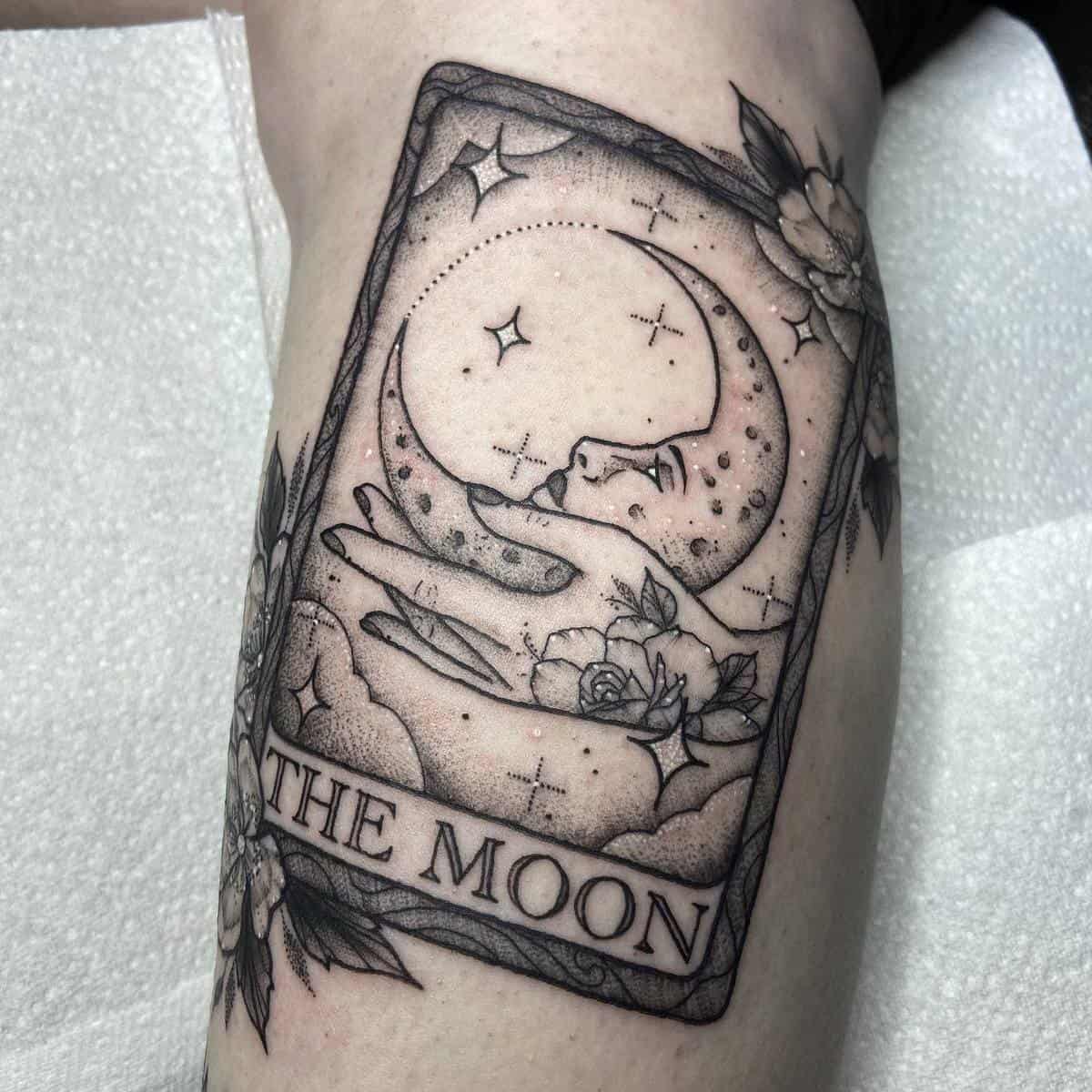 A tattoo of a card with the moon, stars, flowers, and the text "THE MOON" on an arm