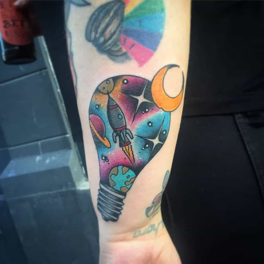 Tattoo on arm: a light bulb with a space scene including a rocket, planets, and stars