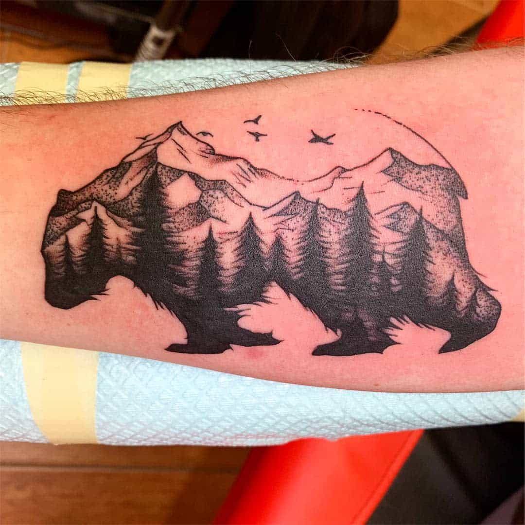 Tattoo of a bear silhouette with a mountain, forest, and birds inside