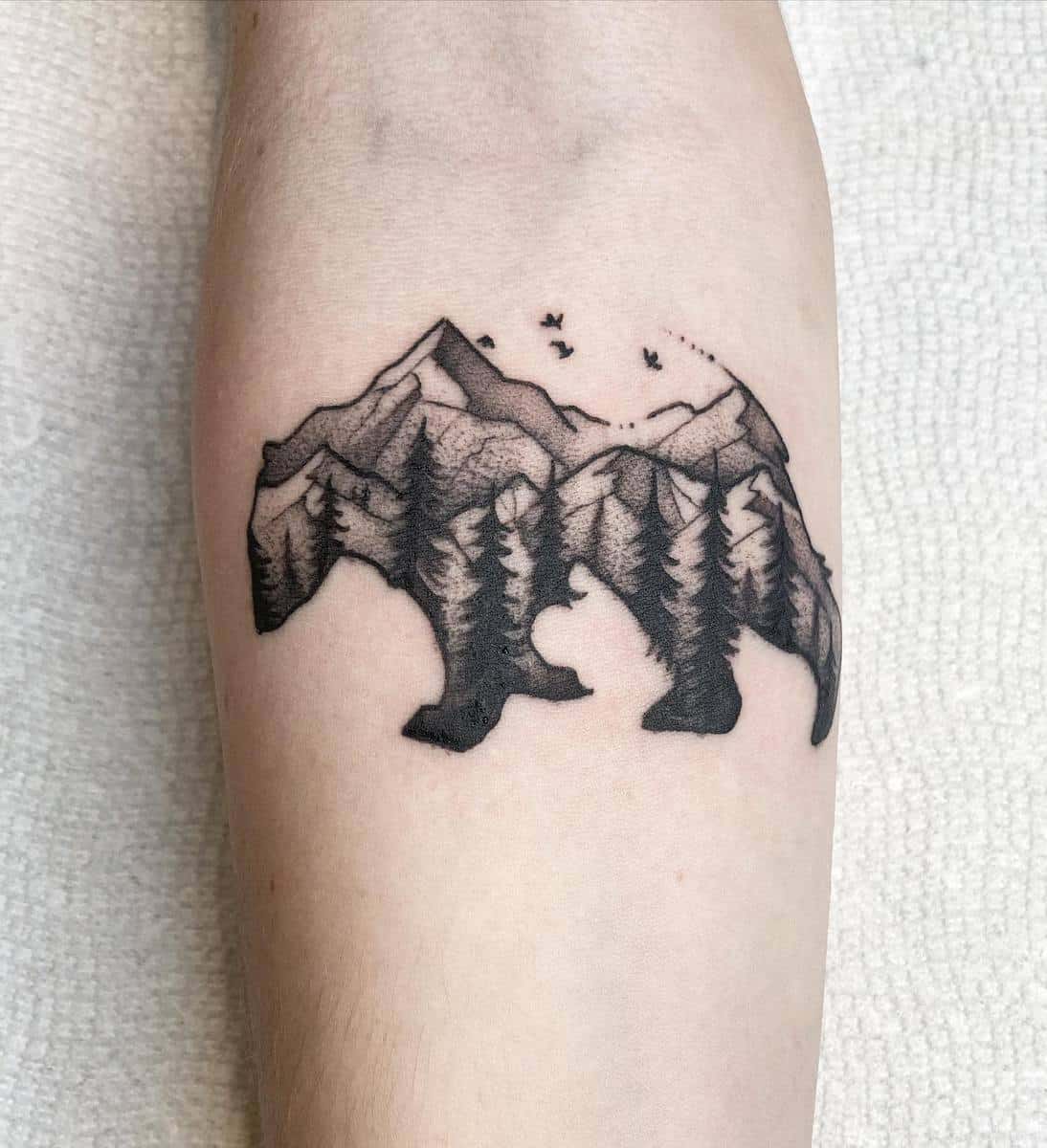Tattoo of a bear silhouette with mountains, trees, and birds on an arm