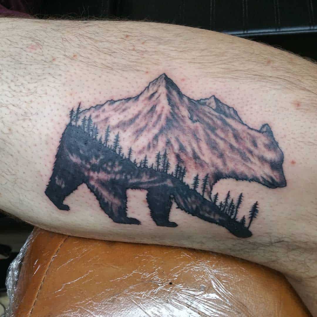 Tattoo of a bear silhouette showcasing a serene mountain scene with trees nestled inside it on a person's arm