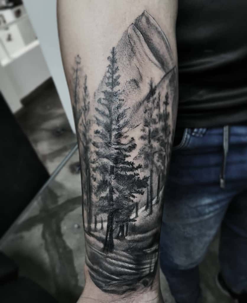 Tattoo on a forearm depicting a forest with tall trees and a mountain in the background