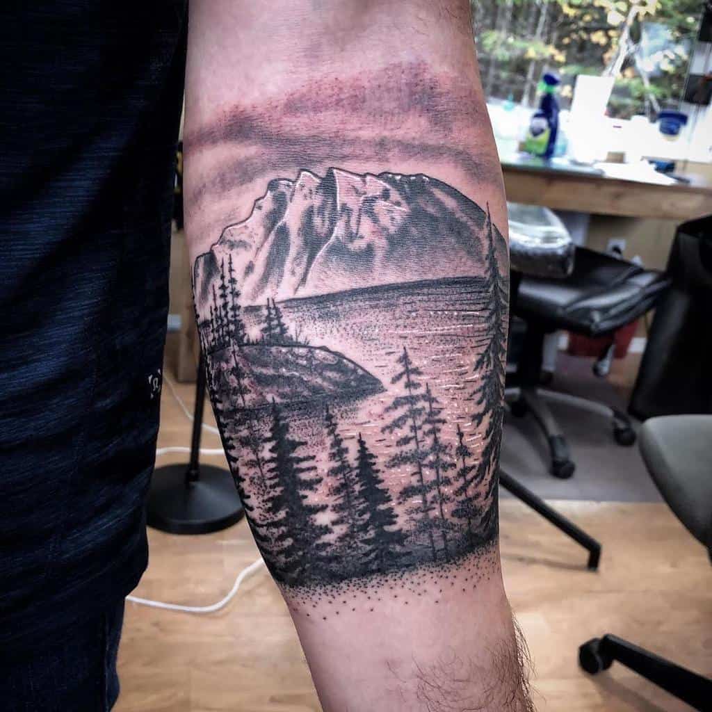 Tattoo depicting a mountain landscape with trees and a body of water, located on a person's forearm