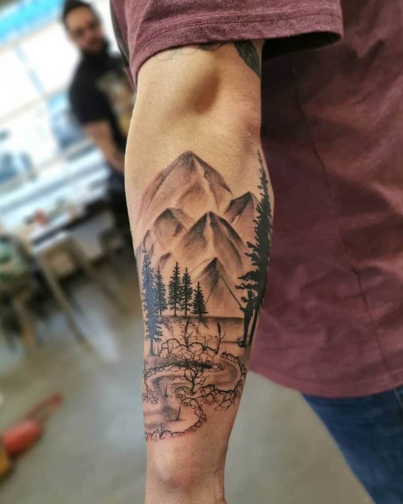 Forearm tattoo of mountains, silhouetted trees, and a winding river in a realistic black and gray style